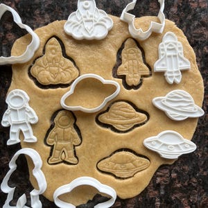 Puede incluir: Una lámina de masa para galletas con varios cortadores de galletas con temática espacial. Los cortadores son de plástico blanco e incluyen formas de cohetes, astronautas, planetas, nubes y ovnis. La masa es de color marrón claro, con las formas recortadas visibles.