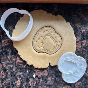 Könnte beinhalten: Ein weißer Plastik-Ausstecher in Form von zwei sich überlappenden Kreisen. Der Ausstecher liegt auf einer braunen Arbeitsfläche neben einem runden Stück Keksteig mit der Abdruck des Ausstechers.