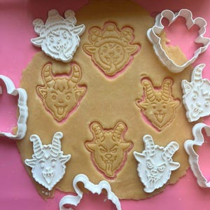 May include: A collection of white plastic cookie cutters shaped like various horned goat heads, some of which are pressed into a flat piece of light brown cookie dough, creating detailed impressions of faces with horns, tongues, and expressive features.