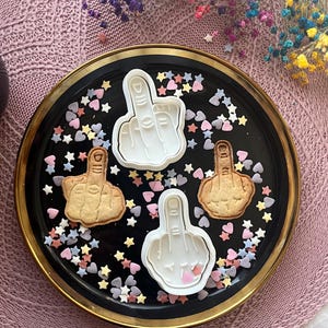 May include: A black plate with a gold rim holds four cookies and cookie cutters shaped like a hand giving the middle finger. The cookies are tan, and the cutters are white. Colorful sprinkles surround the cookies.