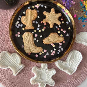 May include: A black plate with a gold rim holds cowboy-themed cookies: a cactus, sheriff's star, cowboy boot, and hat. White cookie cutters in the same shapes are nearby. Colorful star and heart sprinkles add a festive touch. A candle and flowers are in the background.