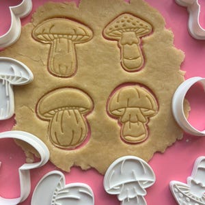 Può includere: Un set di tagliabiscotti in plastica bianca a forma di fungo disposti attorno a un pezzo di pasta frolla stesa. L'impasto presenta quattro distinte impronte di funghi, ognuna con diverse texture del cappello e dettagli del gambo.