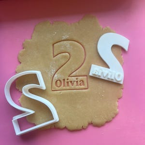 Può includere: Un pezzo di pasta con il numero 2 e il nome "Olivia" impresso. Due formine per biscotti in plastica bianca a forma di lettera "S" sono accanto alla pasta. La pasta è su una superficie rosa.