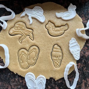 Puede incluir: Primer plano de masa para galletas con varios cortadores de galletas con temática de verano. Los cortadores son de plástico blanco e incluyen formas como un sombrero para el sol, chanclas, un cono de helado y una concha. La masa es de color marrón claro.