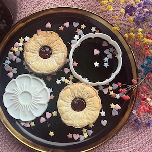 May include: A black plate with a gold rim displays two jam biscuits, a white flower-shaped mould, and a white scalloped-edge mould. Heart and star sprinkles in various colours surround the biscuits. The biscuits have a flower-like design.