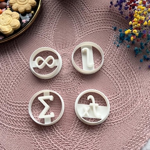 Op de afbeelding: Vier witte plastic koekjesvormen met verschillende wiskundige symbolen: oneindigheid, integraal, sigma en een breuk. De vormen liggen op een roze gehaakte placemat.