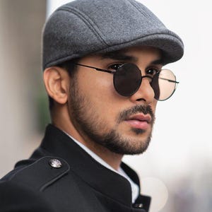 Puede incluir: Un hombre con una gorra de golf gris y gafas redondas negras. Lleva una chaqueta negra con cuello mandarín.