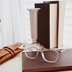 May include: A brown leather glasses case with the text "GOYA" and "HANDCRAFTED IN ITALY" embossed on the front. The case is lying on a white surface next to a pair of clear glasses and a brown leather-bound journal.