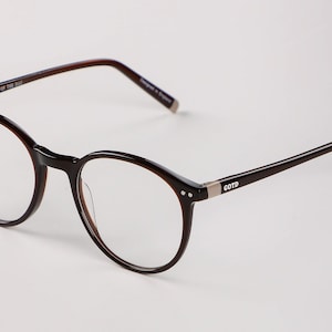 May include: Dark brown eyeglasses with a round frame. The glasses feature a thin frame and temples, with the text "GLASSES OF THE DAY" on the top and "GOTD" on the temple. The lenses are clear.