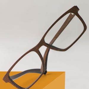May include: Brown wooden frame spectacles with clear lenses. The glasses are resting on a yellow surface.