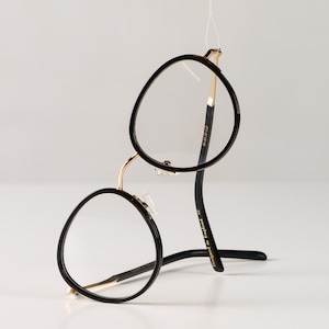 Puede incluir: Gafas redondas negras y doradas con un fino marco dorado y patillas de plástico negro. Las patillas están marcadas con "Designed in England."