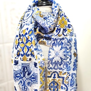 May include: A white scarf with a blue and yellow tile pattern, displayed on a mannequin. The scarf has a repeating design of floral and geometric shapes. The edges are finished with small yellow tassels.