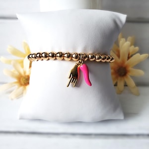 May include: Gold beaded bracelet with a gold hand charm and a pink chili pepper charm. The bracelet is displayed on a white pillow with yellow flowers in the background. The bracelet is a fashion accessory.
