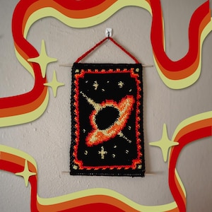May include: A black and orange crocheted wall hanging depicting a black hole with a bright orange accretion disk and stars. The hanging is framed with a red and orange border.