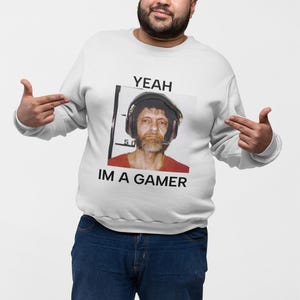 May include: A white sweatshirt with the text "YEAH IM A GAMER" and a mugshot-style image of a man wearing headphones. The person in the image is wearing blue jeans and pointing at the text on the sweatshirt.