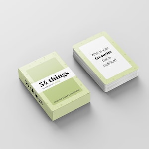 May include: A light green box and a stack of cards with the text "54 things to ask your family". The cards have a light green border and the question "What is your favourite family tradition?" printed on them. The box and cards are on a light gray surface.