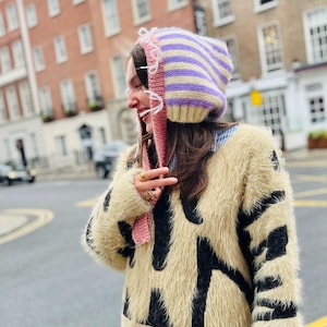 May include: A striped knit hat in lavender, cream, and yellow with pink ear flaps. The hat is paired with a cream-colored fuzzy sweater with black abstract shapes. The person is wearing a blue and white striped shirt underneath.