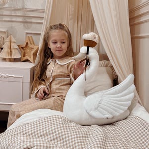 Puede incluir: Una niña con pijama a cuadros beige está sentada en una cama con una gran almohada blanca con forma de cisne. La almohada tiene un sombrero marrón y una corbata negra. Una decoración de madera en forma de estrella y árbol está en una mesita de noche.