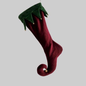 May include: A burgundy velvet Christmas stocking with a green velvet cuff. The cuff has a jagged edge and is adorned with small gold-coloured bells. The stocking has a curled toe and a gold bell at the tip.