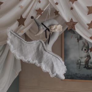 Pode incluir: Móvel de cisne cinzento com asas com bordas de renda e um chapéu extravagante, suspenso de um dossel adornado com decorações de estrelas douradas. O cisne tem uma fita preta e uma gola com babados creme. Uma imagem emoldurada é visível no fundo.