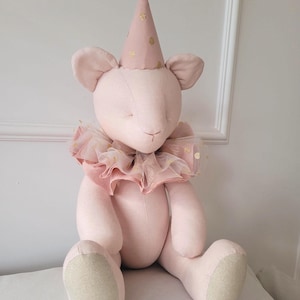Può includere: Una bambola di topo rosa di peluche con un cappello da festa a pois dorati e un colletto a balze. La bambola ha grandi orecchie ed è seduta. I piedi della bambola sono di colore beige chiaro.