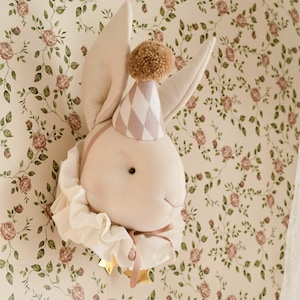 Puede incluir: Un adorno de pared con forma de cabeza de conejo de peluche, con un gorro de fiesta y un cuello con volantes. El gorro tiene un estampado de rombos rosas y blancos y un pompón marrón. Fondo de papel pintado floral.
