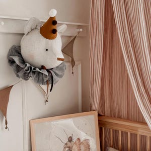 Pode incluir: Decoração de parede com cabeça de elefante de pelúcia. O elefante é branco com orelhas cinzentas, um chapéu de festa castanho e dourado e uma gola com babados listrados. Uma faixa castanha pende por baixo e uma imagem emoldurada de uma traça é visível.