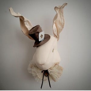 Puede incluir: Decoración de pared con forma de cabeza de conejo blanco roto. Presenta largas orejas caídas, un pequeño sombrero de copa marrón con un símbolo de pica y un cuello con volantes. Una cinta negra está atada en la base del cuello.