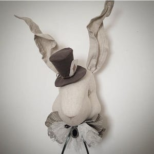 Puede incluir: Cabeza de conejo de tela beige claro, adornada con largas orejas caídas y un pequeño sombrero de copa marrón con un detalle de carta de juego. El conejo también lleva un cuello con volantes. Ideal para la decoración infantil.