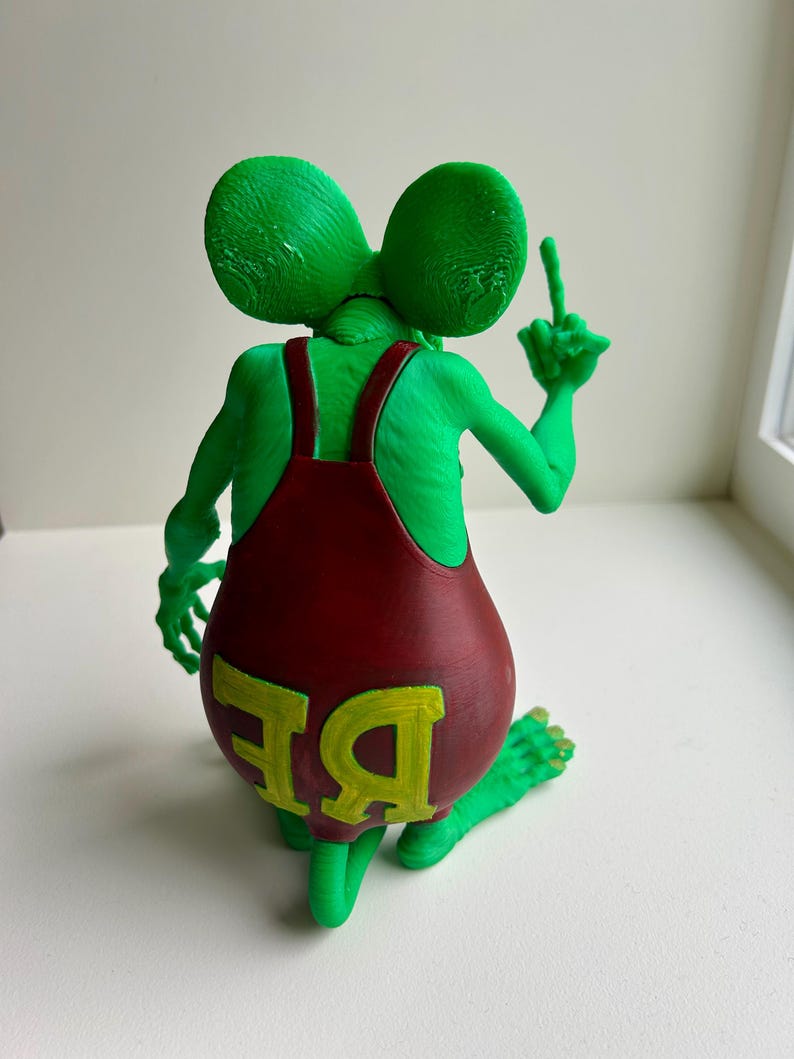 Peut inclure: Figurine verte repr&eacute;sentant une cr&eacute;ature ressemblant &agrave; une souris, v&ecirc;tue d'une salopette rouge avec les lettres "BE" en jaune. La cr&eacute;ature fait un doigt d'honneur. La figurine a de grandes oreilles et est assise.