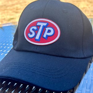 Peut inclure: Casquette de baseball noire avec le logo STP ovale rouge, blanc et bleu sur le devant. La casquette a une visière incurvée et une couronne structurée, idéale pour un usage quotidien.