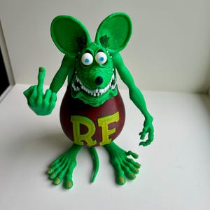 Peut inclure: Figurine de rat vert de dessin anim&eacute; avec de grandes oreilles, des yeux bleus et un large sourire, faisant un doigt d'honneur. Le corps du rat est rouge avec les lettres "RE" en jaune.