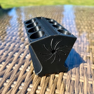 以下が含まれることがあります： 上部に複数の円形開口部がある黒色の 3D プリントオーガナイザー。 オーガナイザーは、側面に様式化されたスパイラルを備えた幾何学的なデザインです。 このオブジェクトはマットブラックの素材でできており、小さなアイテムを収納するように設計されています。