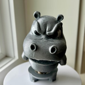 May include: A grey, 3D-printed hippo figurine with an angry expression. The hippo has large eyes, small ears, and a wide mouth. The figurine is sitting on a white, round surface.