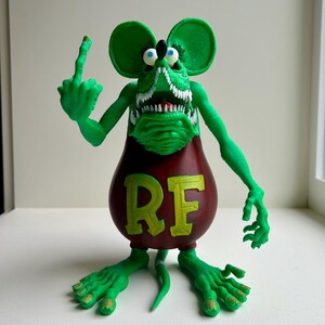 Peut inclure: Figurine de rat vert de dessin anim&eacute; avec de grandes oreilles et une expression mena&ccedil;ante, faisant un doigt d'honneur. Le rat a un corps rouge avec les lettres "RF" en jaune. La figurine est sur une surface blanche.