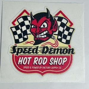 Peut inclure: Un autocollant découpé représentant un personnage de diable rouge avec un cigare, flanqué de deux drapeaux à damier noir et blanc. Le texte indique Speed Demon HOT ROD SHOP et SPEED & POWER BY FACTORY SUPPLY CO. en lettres noires et blanches.