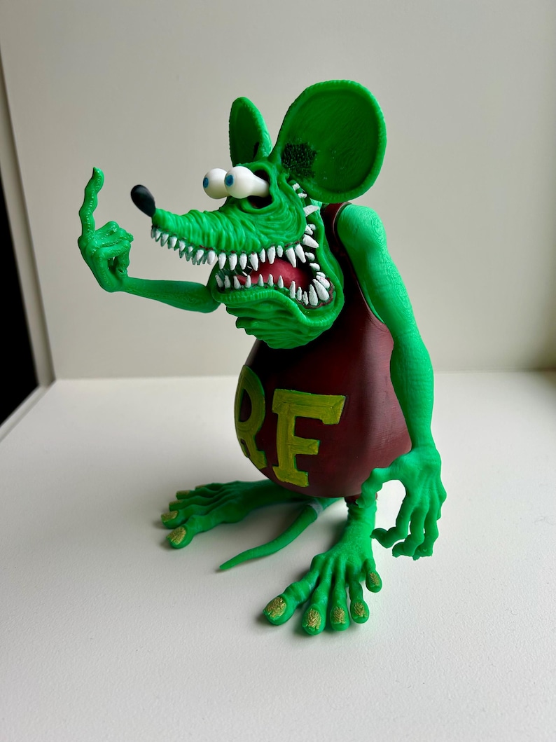 Peut inclure: Figurine de rat de dessin anim&eacute; vert avec de grandes oreilles, des dents blanches et des yeux globuleux. Le rat porte un d&eacute;bardeur bordeaux avec les lettres "RE" en jaune. Le rat fait un doigt d'honneur.