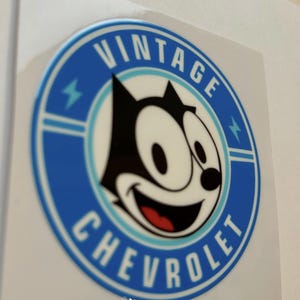 May include: A circular sticker with a blue and white design. It features Felix the Cat and the words "VINTAGE CHEVROLET". The cartoon cat has a black head and a smiling face.