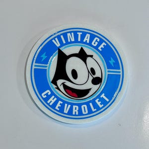 May include: Circular coaster with a blue and white design. The text "VINTAGE CHEVROLET" surrounds a cartoon cat with a black body and white face. The coaster has two blue lightning bolt accents.