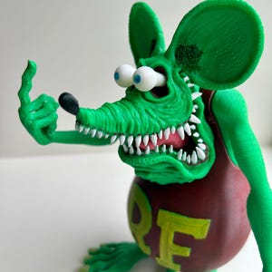 Peut inclure: Figurine de rat vert de dessin anim&eacute; avec de grandes oreilles, des dents blanches et des yeux globuleux. Le rat porte une chemise marron avec le mot "ORE" en jaune. Le rat fait un doigt d'honneur avec sa main gauche.