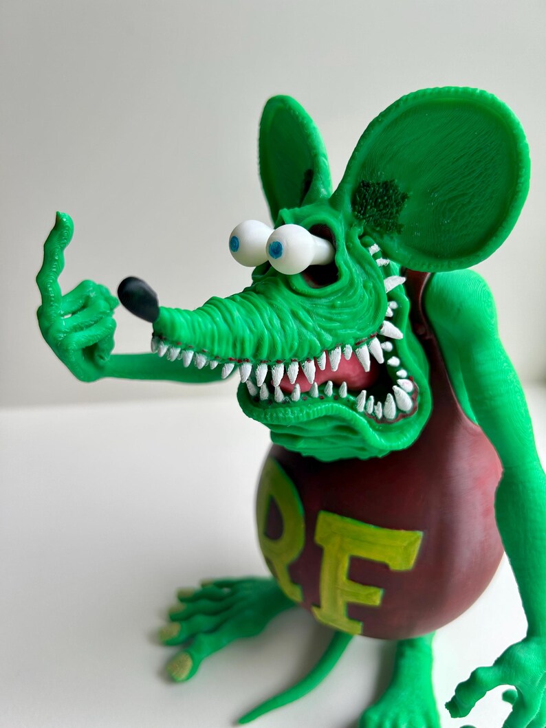 Peut inclure: Figurine de rat vert de dessin anim&eacute; avec de grandes oreilles, des dents blanches et des yeux globuleux. Le rat porte une chemise marron avec le mot "ORE" en jaune. Le rat fait un doigt d'honneur avec sa main gauche.