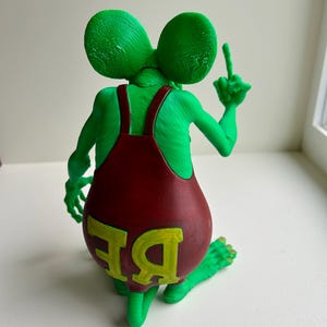 Peut inclure: Figurine verte repr&eacute;sentant un personnage ressemblant &agrave; une souris, portant une salopette rouge avec les lettres "BE" en jaune. Le personnage fait un doigt d'honneur. Il a de grandes oreilles et est &agrave; genoux.