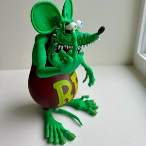 Peut inclure: Figurine de rat vert de dessin anim&eacute; avec un torse rouge et la lettre "R" en jaune. Le rat a de grandes oreilles, un long museau et fait un doigt d'honneur. La figurine a des dents blanches et des yeux bleus.