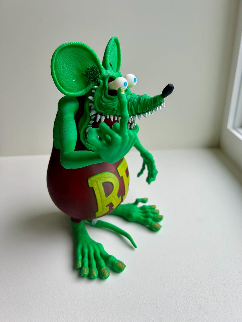 Peut inclure: Figurine de rat vert de dessin anim&eacute; avec un torse rouge et la lettre "R" en jaune. Le rat a de grandes oreilles, un long museau et fait un doigt d'honneur. La figurine a des dents blanches et des yeux bleus.