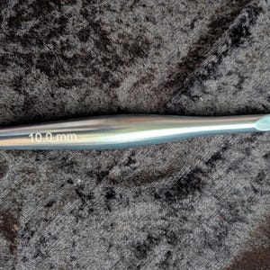 May include: A silver crochet hook with a 10.0 mm size marking. The hook is cylindrical with a rounded tip and a hook at the end. The text "10.0 mm" is etched on the side of the hook.