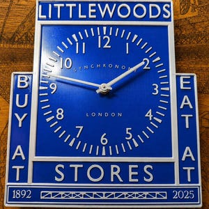 May include: A rectangular blue and white wall clock featuring the text LITTLEWOODS at the top, BUY AT on the left, EAT AT on the right, and STORES at the bottom. The clock face displays the text SYNCHRONOME LONDON, numbers 1-12, and 1892-2025.
