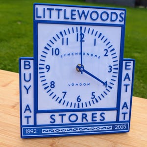 Puede incluir: Un reloj azul y blanco con las palabras "LITTLEWOODS", "BUY AT STORES" y "EAT AT" en letras azules. La esfera del reloj muestra la hora, con la palabra "SYNCHRONOME" y "LONDON" visibles. El reloj está sobre una superficie de madera.