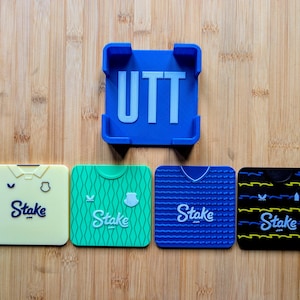 Puede incluir: Un soporte azul para teléfono con las letras "UTT" y cuatro posavasos cuadrados. Los posavasos presentan diseños de camisetas en amarillo, verde, azul y negro, cada uno con el logotipo "Stake". Los artículos están dispuestos sobre una superficie de madera.