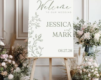 May include: A wedding welcome sign on a wooden easel. The sign is cream-colored with green floral accents and text that reads "Welcome to our Wedding" and the names "Jessica and Mark" with the date "08.27.28".