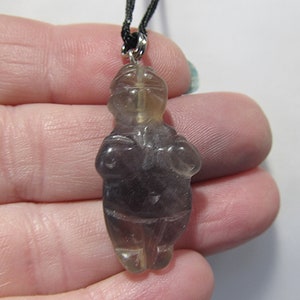 May include: A purple and clear crystal pendant carved in the shape of a human figure. The pendant is hanging from a black cord.